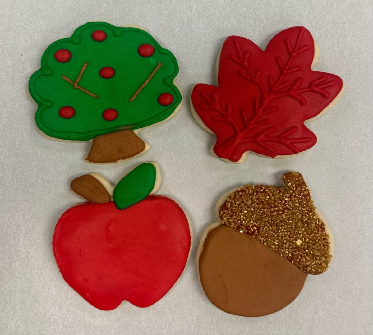 Four decorated sugar cookies shaped like a green tree, a red maple leaf, a red apple, and a brown acorn with a glittery gold cap, arranged on a white background.